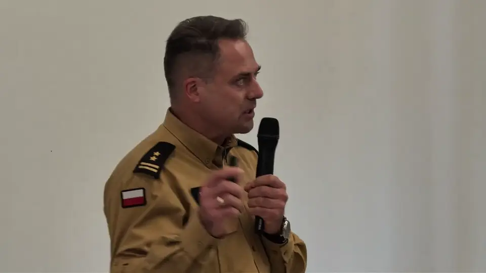 Strażak w mundurze mówi do mikrofonu — kadr z nagrania spotkania. Otwiera nagranie w YouTube.
